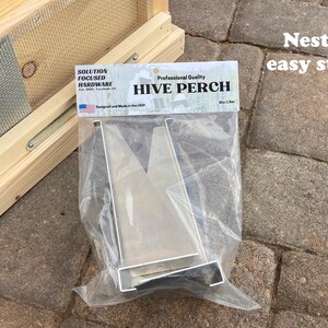 Op de afbeelding: Een zilveren metalen bijenkorf-zitstok voor imkerij. De zitstok is ontworpen om te worden gebruikt met een bijenkorf en is gemaakt van duurzaam metaal. De verpakking vermeldt "Professional Quality Hive Perch".