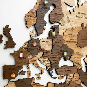 Könnte beinhalten: Eine Holzkarte von Europa mit Umrissen der Länder in verschiedenen Brauntönen. Kleine Pins mit Flaggen markieren Standorte. Die Karte umfasst Länder wie Frankreich, Deutschland und Italien. Die Karte ist auf einem weißen Hintergrund montiert.