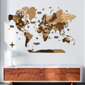 Può includere: Una mappa del mondo in legno nei toni del marrone, con i contorni dei paesi e le etichette degli oceani. Sono inclusi aerei e una bussola decorativi. La mappa è montata su una parete bianca sopra un mobile in legno.