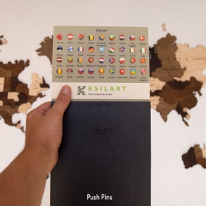 May include: A close-up of a black card with the text "Push Pins" and the logo "KSILART laser engraving studio". The card is being held by a hand in front of a wooden world map with small flags representing different countries.