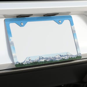 Peut inclure: Un cadre de plaque d'immatriculation bleu et blanc avec un motif de montagne et de forêt. Le cadre est monté à l'arrière d'une voiture blanche. Le motif présente des montagnes enneigées, des arbres verts et un ciel bleu.