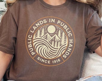May include: A brown t-shirt featuring a vintage-style graphic of a mountain landscape and the text "Public Lands in Public Hands Since 1916".