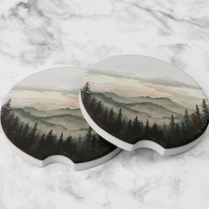 May include: Two car coasters with a forest and mountain landscape design. The coasters are round with a small notch and a white border. The artwork features a misty mountain scene with evergreen trees in shades of green and brown.