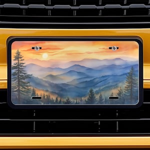 May include: A license plate featuring a scenic mountain landscape with a sunset. The plate is mounted on a yellow vehicle, with a black grill and the text "RELAXING JOURNEYS" above the plate.