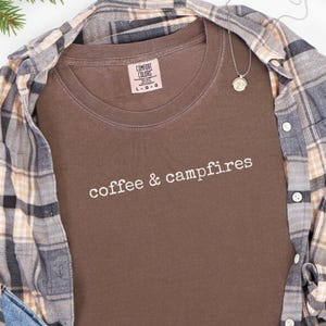 Peut inclure: Un t-shirt marron avec l'inscription "coffee & campfires" en lettres blanches. Le t-shirt est associé à une chemise à carreaux, un jean bleu et des bottes marron. Un collier argenté avec un pendentif est également visible.