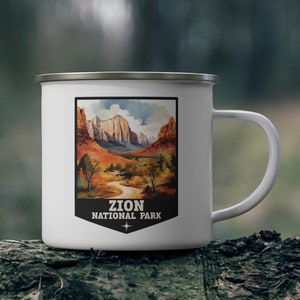 May include: White enamel mug featuring a landscape illustration of Zion National Park. The artwork depicts a scenic view of the park with mountains, a river, and trees. The mug has a silver rim and a white handle, with the text "ZION NATIONAL PARK".