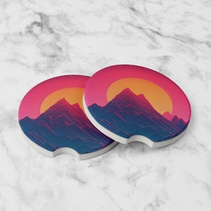 May include: Two round pink and blue coasters with a retro mountain and sunset design. The coasters are made of ceramic and have a cork backing.