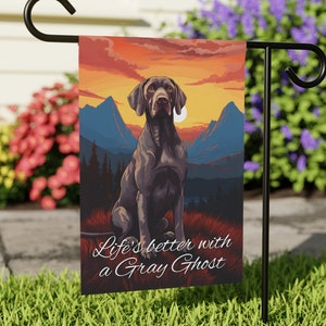 May include: A garden flag with a brown and black German Shorthaired Pointer dog sitting in front of a mountain landscape with a sunset sky. The text on the flag reads "Life's better with a Gray Ghost".