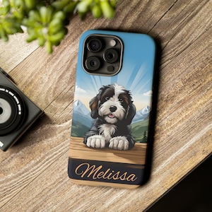 May include: A blue and white phone case with a cartoon dog in front of a mountain scene. The dog is black and white and has a white chest. The case has the name "Melissa" printed on the bottom.