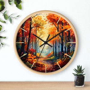 May include: A circular wall clock with a light wood frame. The clock face depicts a vibrant autumn forest scene with a winding path, trees, and falling leaves in orange, red, and yellow hues. Black clock hands.