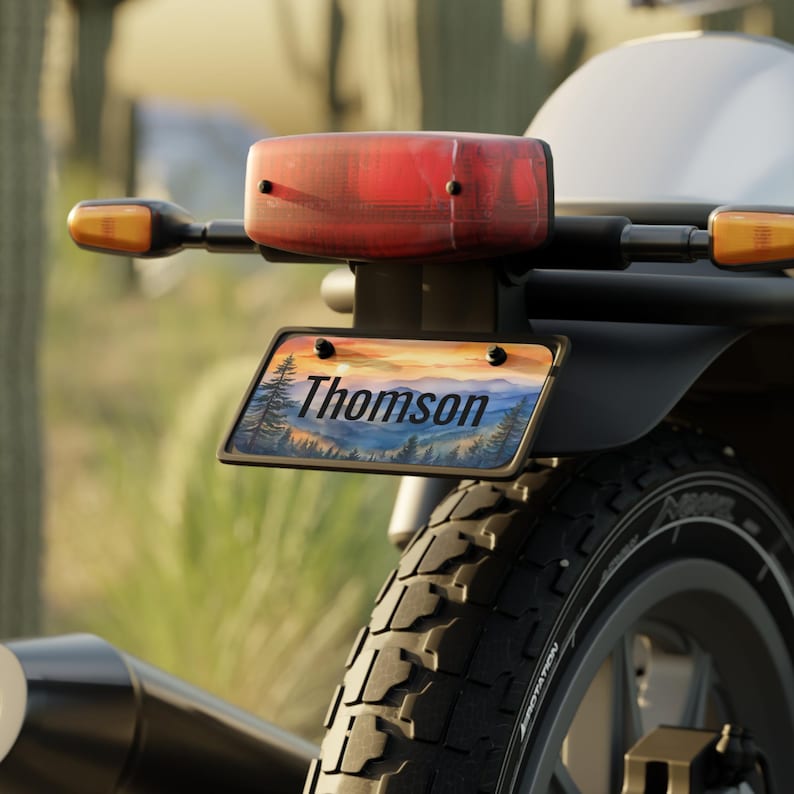 May include: Motorcycle license plate with the name "Thomson" and a scenic landscape of mountains and trees. The plate is mounted on the rear of a motorcycle, with a red taillight and amber turn signals visible.