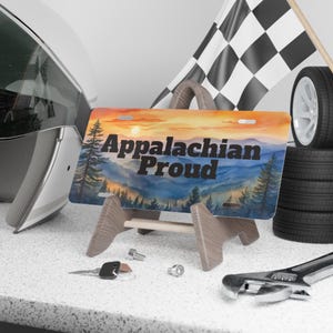 May include: License plate with the text "Appalachian Proud" featuring a watercolor landscape of mountains and trees under a sunset sky. The plate is displayed on a wooden stand, with a racing flag and tires in the background.