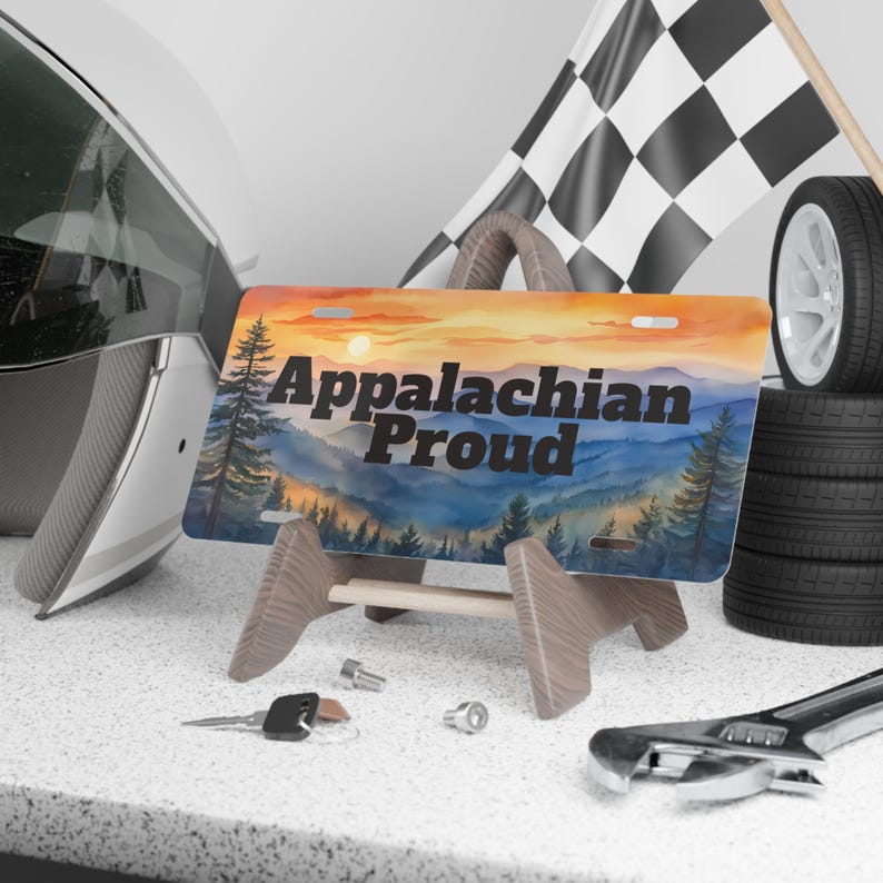 May include: License plate with the text "Appalachian Proud" featuring a watercolor landscape of mountains and trees under a sunset sky. The plate is displayed on a wooden stand, with a racing flag and tires in the background.