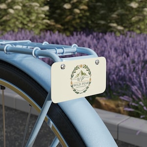 May include: A light blue bicycle with a cream-colored license plate attached to the rear. The plate features a mountain logo with the text "Wild Peak Photography" in a circular design. The background includes purple flowers.