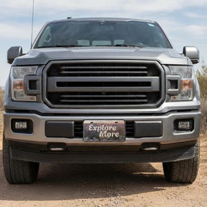 May include: Front view of a gray pickup truck with a black grille and bumper. The license plate reads "Explore More." The truck is parked on a dirt road with a blue sky in the background. The truck has large tires and headlights.