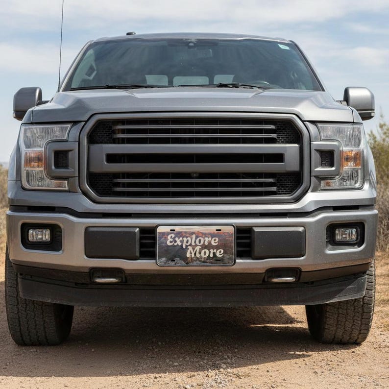 May include: Front view of a gray pickup truck with a black grille and bumper. The license plate reads "Explore More." The truck is parked on a dirt road with a blue sky in the background. The truck has large tires and headlights.