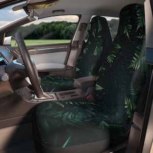 May include: Car seat covers with a dark green fern leaf pattern on a black background. The interior of a car is visible, including the steering wheel, dashboard, and a view of the outside landscape through the window.