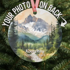 May include: A round, white ornament with a watercolor painting of a mountain range and forest. The text "Kotenay National Park 2023" is printed on the ornament.