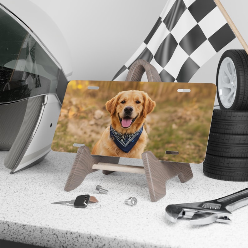 May include: A license plate featuring a Golden Retriever wearing a blue bandana, displayed on a wooden stand. A racing flag, tires, and tools are in the background, suggesting a car-themed product.