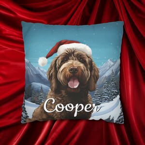 May include: Square decorative pillow featuring a brown dog wearing a Santa hat, with the name "Cooper" in white script. The background shows a snowy mountain scene with pine trees and a blue sky. The pillow is likely made of soft fabric.