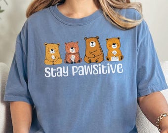 May include: A blue t-shirt featuring a graphic of four brown bears sitting in a row. The text "Stay Pawsitive" is printed below the bears.