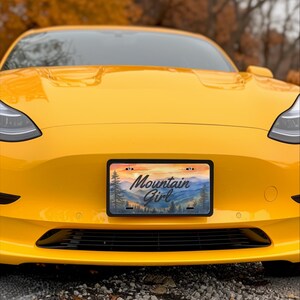May include: A yellow car with a license plate that reads "Mountain Girl." The license plate features a watercolor landscape of mountains and trees under a sunset sky. The car's front end is visible.