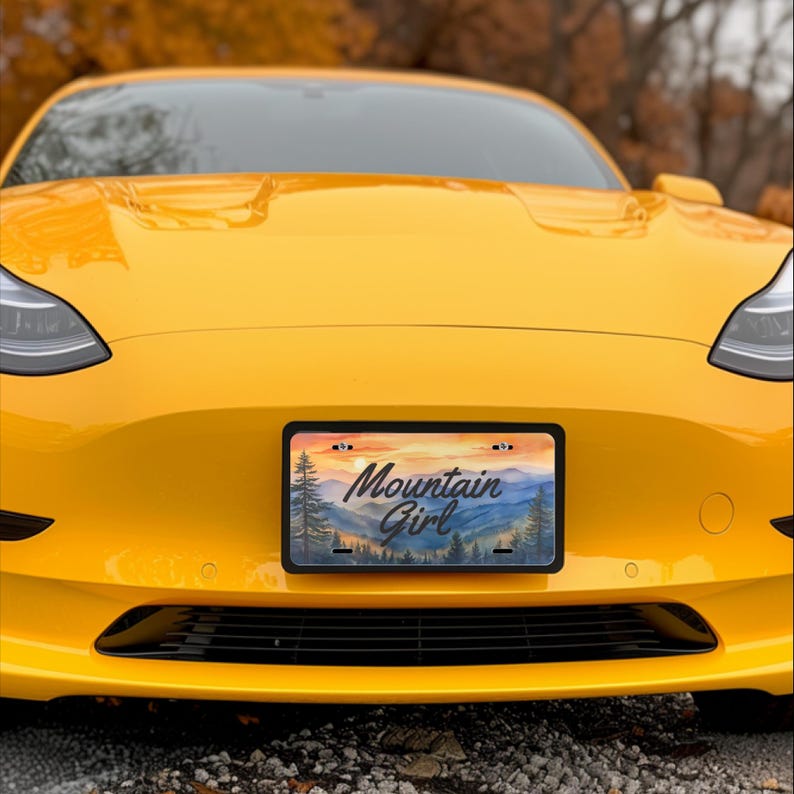 May include: A yellow car with a license plate that reads "Mountain Girl." The license plate features a watercolor landscape of mountains and trees under a sunset sky. The car's front end is visible.