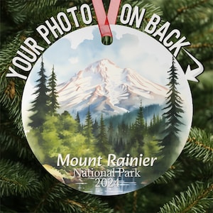 May include: A round, white, Christmas ornament with a watercolor painting of Mount Rainier National Park. The mountain is snow-capped and surrounded by green trees. The text "Mount Rainier National Park 2024" is printed on the ornament.
