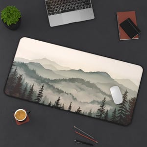 May include: A large desk mat with a watercolor design of a misty mountain range with a forest in the foreground. The mat is on a black surface with a laptop, a cup of coffee, pencils, and a mouse.