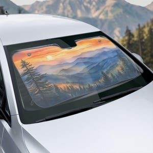 May include: A car windscreen sunshade with a watercolour landscape of mountains and a sunset. The artwork shows layers of blue and purple mountains under an orange and yellow sky, with silhouetted trees in the foreground.