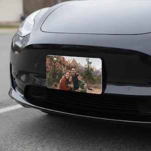 May include: A rectangular license plate featuring a family portrait against a backdrop of mountains and trees. The plate is mounted on the front of a black car. The family is smiling and hugging each other.