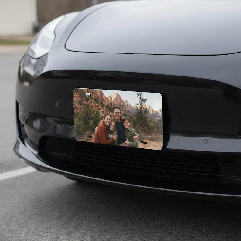 May include: A rectangular license plate featuring a family portrait against a backdrop of mountains and trees. The plate is mounted on the front of a black car. The family is smiling and hugging each other.