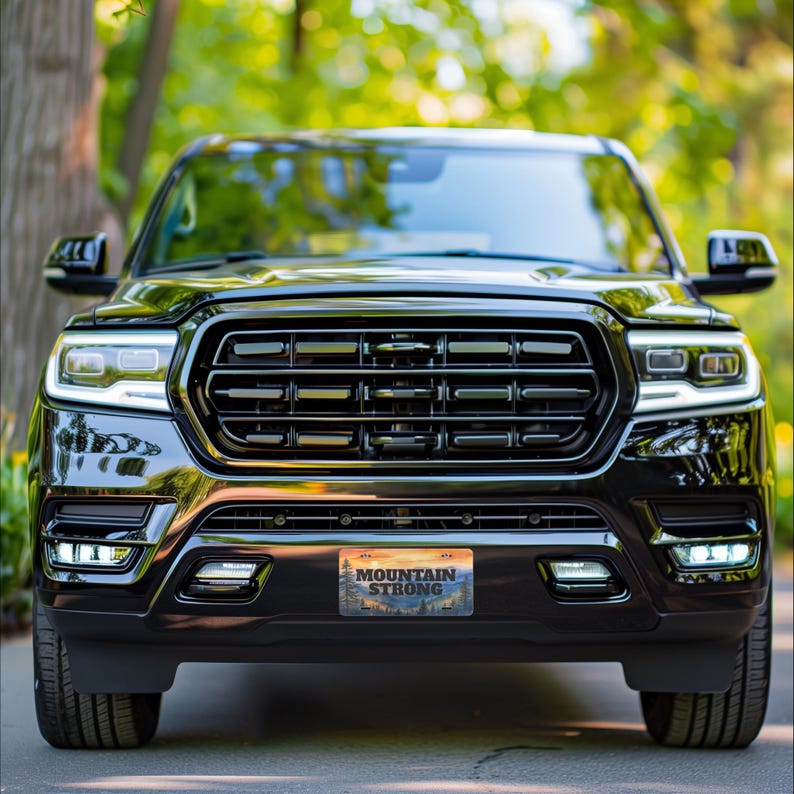 May include: A black SUV with a prominent front grille and bright headlights. The license plate reads "MOUNTAIN STRONG" with a scenic mountain and sunset design. The vehicle is parked in front of a blurred green background of trees.