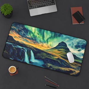 May include: Rectangular mouse pad featuring a landscape with a waterfall, mountain, and aurora borealis. The design includes shades of blue, green, yellow, and orange. A white computer mouse is on the pad. A laptop, notebook, and pencils are also visible.