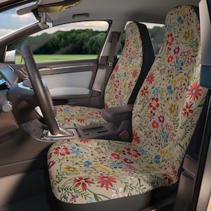 May include: Car seat covers featuring a floral pattern with red, blue, yellow, and white flowers on a tan background. The covers are installed in a car, with the steering wheel and dashboard visible.