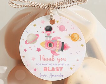 Pode incluir: Etiqueta redonda de agradecimento rosa e dourada com um astronauta em um traje espacial rosa montando um foguete rosa com estrelas e planetas ao fundo. O texto na etiqueta diz "Obrigado por fazer da minha festa uma explosão, com amor, Amanda."