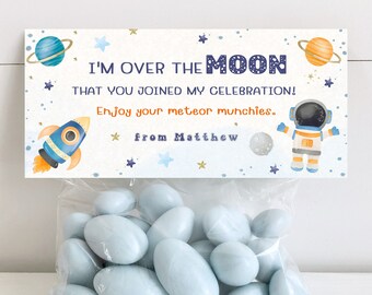 May include: A blue and white favor bag with a space-themed tag that reads "I'm over the moon that you joined my celebration! Enjoy your meteor munchies. from Matthew". The tag features a rocket, astronaut, planets, and stars.