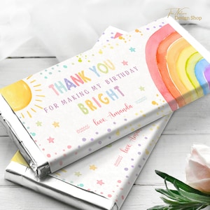 Puede incluir: Envoltorio de barra de chocolate con temática de arcoíris con el texto "THANK YOU FOR MAKING MY BIRTHDAY BRIGHT" y "love. Amanda". El envoltorio presenta un arcoíris de acuarela, un sol y estrellas sobre un fondo blanco con lunares de colores.