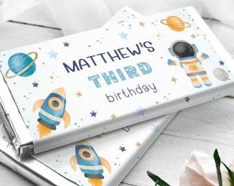 May include: A white chocolate bar wrapper with a blue and orange rocket, a blue planet with a ring, and a gold astronaut. The text on the wrapper says "Matthew's Third birthday".