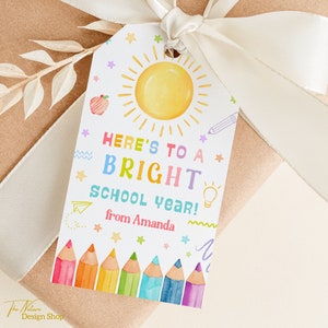 May include: A white gift tag with a colorful illustration of a sun, pencils, and stars. The text reads "Here's to a bright school year! from Amanda".