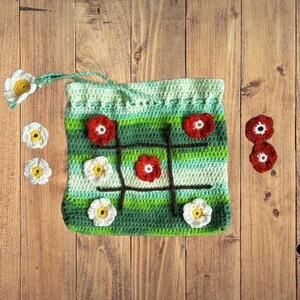 May include: A green and white crocheted bag with a tic-tac-toe board design. Red, white, and green crocheted flowers are attached to the bag.