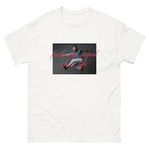 以下が含まれることがあります： 白いTシャツ。空中を飛んでいる人のグラフィックと、ピンクの筆記体で「fuck my stupid shit life」というテキスト。その人は青いシャツと白いズボンを着ています。