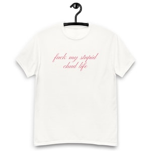 Puede incluir: Camiseta blanca con la frase "fuck my stupid chud life" en escritura cursiva rosa. La camiseta está colgada de una percha negra. La camiseta es una prenda informal.