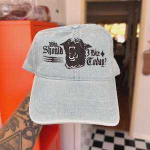 May include: Light blue denim baseball cap with the text "Who Should I Bite Today?" and a black graphic of a snarling dog. The cap has a curved brim and an adjustable strap.