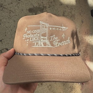 May include: Tan baseball cap with a black and white rope detail. The cap features white embroidered text and a guillotine graphic that reads "NO ONE Protests LIKE The French."