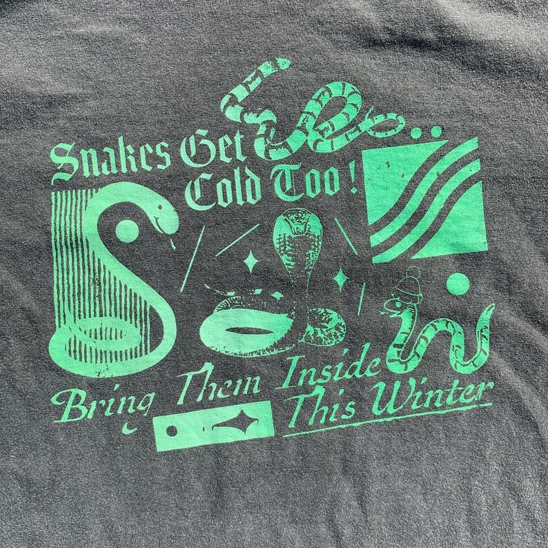 May include: Green graphic print on a dark grey t-shirt featuring snakes and the text "Snakes Get Cold Too! Bring Them Inside This Winter."