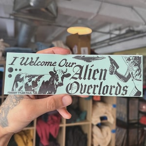 Può includere: Un adesivo rettangolare con sfondo verde chiaro. L'adesivo presenta il testo "I Welcome Our Alien Overlords" in nero, insieme a illustrazioni di una mucca, un UFO e un alieno che tiene un'arma. Il nome dell'artista, "Freddy Tyler Paul", è in basso.