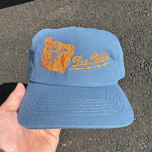 Könnte beinhalten: Eine hellblaue Baseballkappe mit einer gestickten orangefarbenen Bärenkopf-Grafik auf der linken Seite und dem Schriftzug "Da Bears" in orangefarbener Schreibschrift mit einer dekorativen Linie darunter auf der Vorderseite.