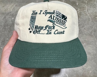 May include: A cream-colored baseball cap with a dark green brim. The front features dark green embroidery with the text "Yea I Speak AUSSIE Now Fuck Off... Ya Cunt" alongside a graphic of a knife and a flag with stars.