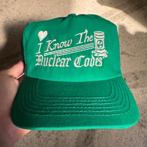 May include: Green baseball cap with white embroidered text and a heart. The text reads "I Know The Nuclear Codes" with a graphic of a nuclear waste container. The cap has a curved brim and a structured crown.